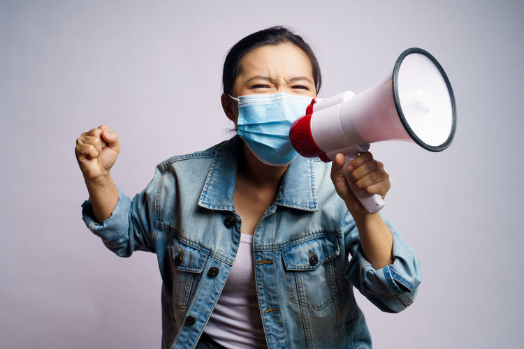 Portrait of young woman wearing flu mask holding megaphone standing against gray background
