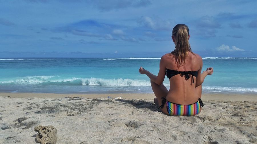 Rear view of woman wearing bikini and meditating on beach against sky
