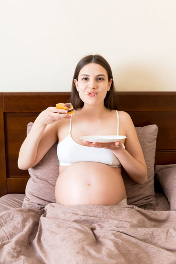 Hungry pregnant woman relaxing in bed is eating greedily a piece of cake and has a dirty mouth.