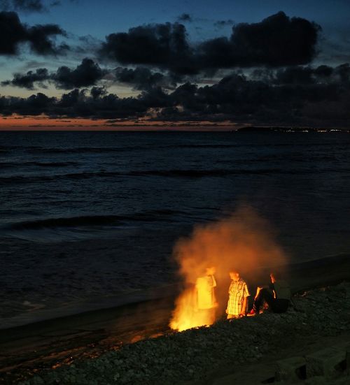 Boys by campfire at beach against cloudy sky during sunset