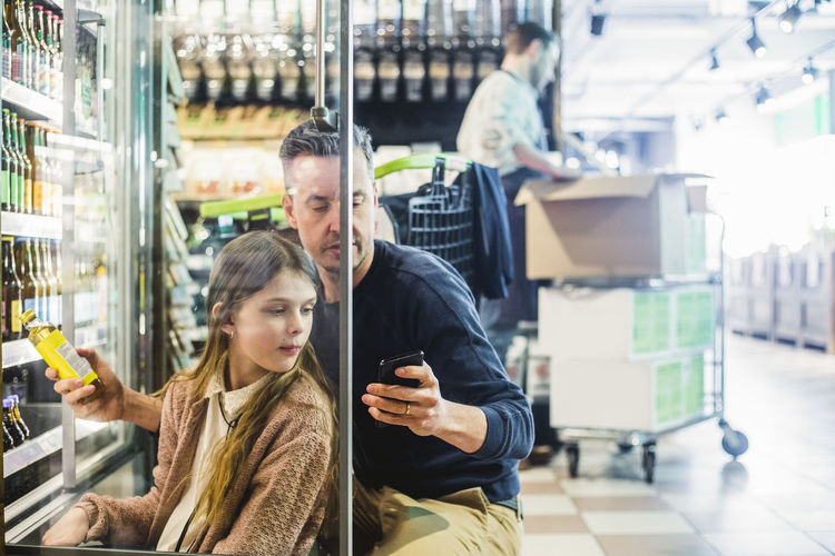 Father and daughter looking at mobile phone while crouching by refrigerator in store