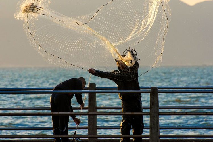 Fishermen working on pier by sea against sky during sunset