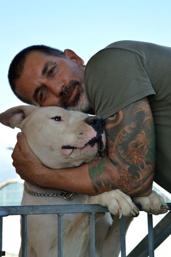 Low angle portrait of tattooed man holding american bulldog against sky