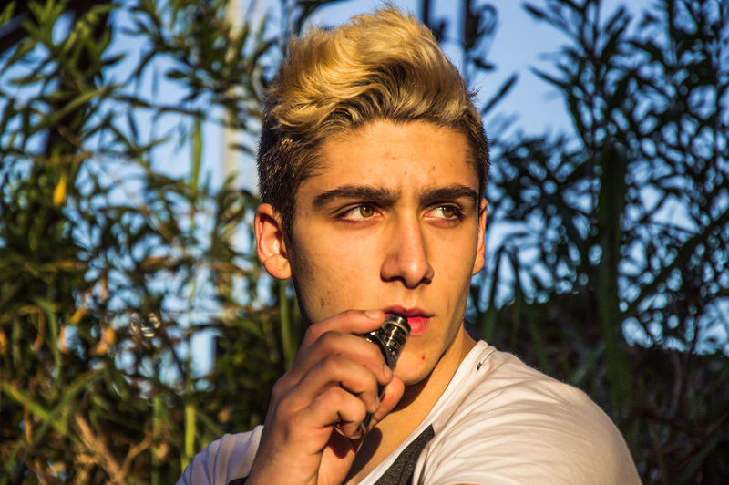 Close-up of teenage boy smoking electronic cigarette against plants