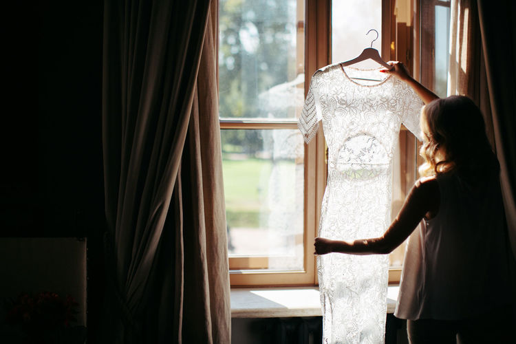 Woman holding and looking at beautiful white wedding dress on a hanger, standing near window at home