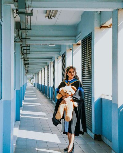 Woman in graduation gown holding teddy bear while standing in corridor
