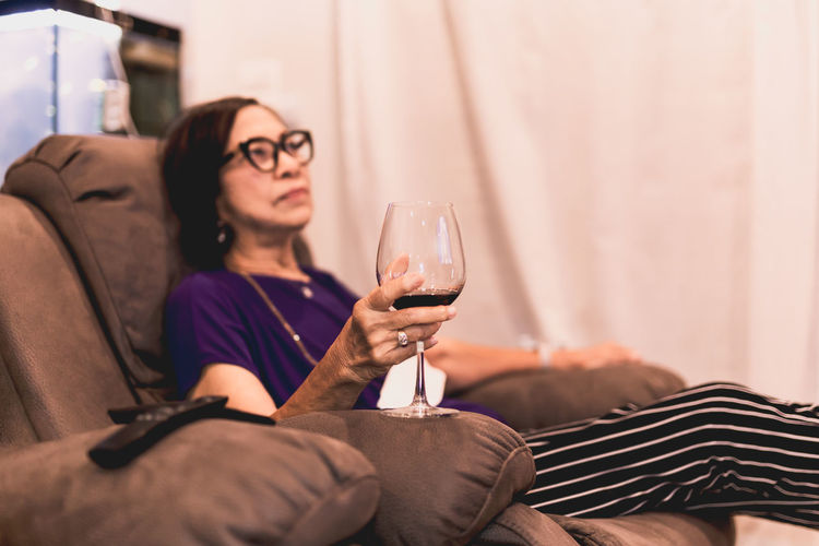 Senior woman sitting on couch having glass of red wine at home.