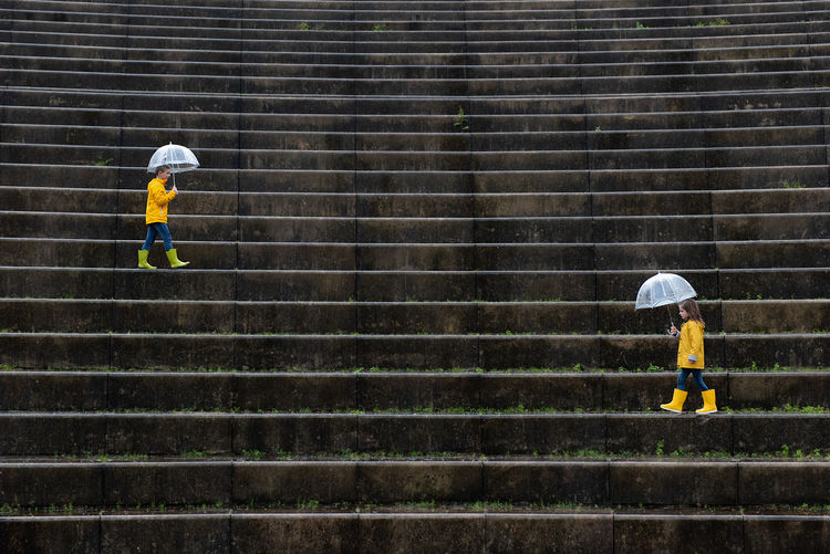 Side view of children in vivid yellow raincoats and rubber boots walking under umbrellas along wet stone stairs in street on rainy day