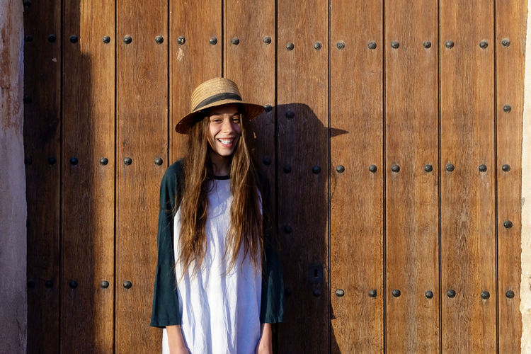 Portrait of young woman wearing hat standing against wooden wall