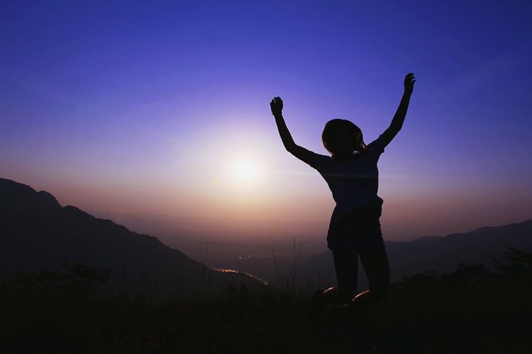 Silhouette woman with arms raised jumping on mountain against sky during sunset
