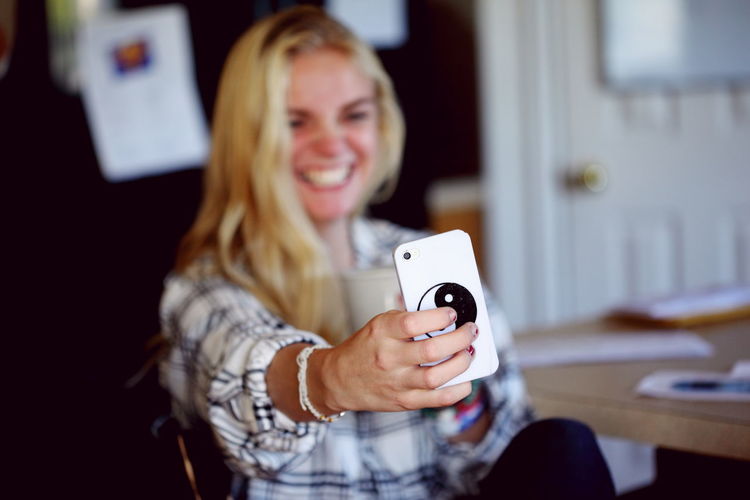 Cheerful young woman taking selfie on mobile phone by table