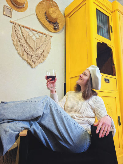 Young woman in beret sits in cozy home, room with glass of wine in hands. vintage yellow cabinet. 