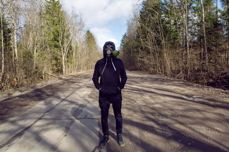 Guy stands in a post apocalyptic forest during the day wearing a gas mask during a coronavirus