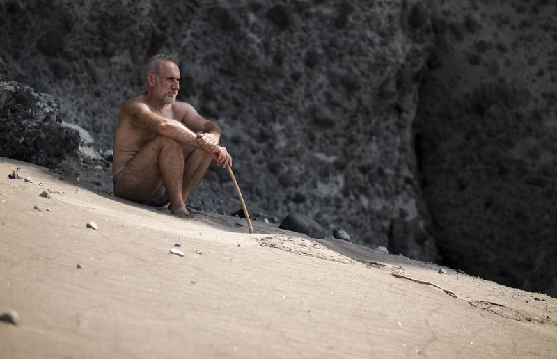Full length of shirtless adult man in swimwear sitting on beach against rock