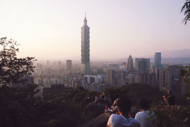 Rear view of people looking at taipei 101 amidst buildings in city during sunset
