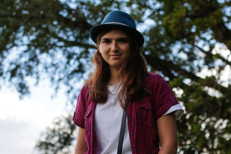 Portrait of a smiling young woman with brown hair wearing a hat outdoors. green blue nature 