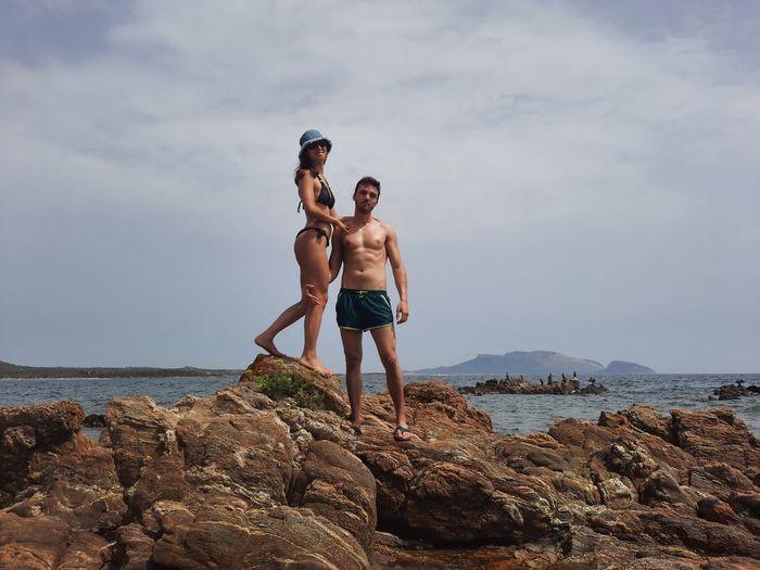 Full length of young couple standing on rock by sea against sky