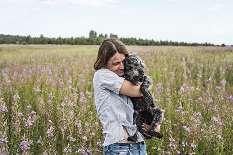 Young smiling woman holding gray fluffy senior dog in hands on field of flowers fireweed , pet love