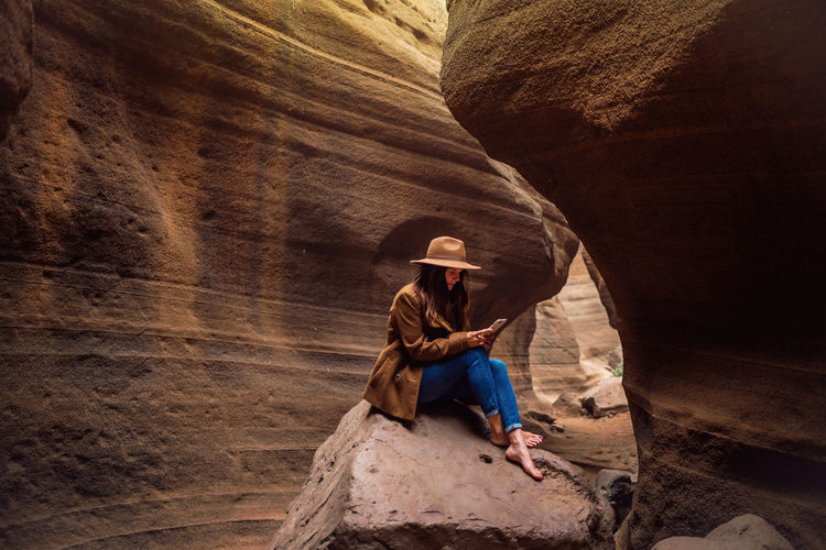 Side view of stylish young barefooted female traveler in hat using smartphone while sitting on stone in rocky ravine and resting after hiking