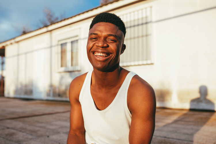 Happy african american male in undershirt with modern haircut looking at camera against building in sunlight