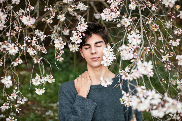 Young male with closed eyes standing under branches with flowers of blooming almond tree in spring garden