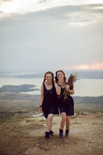 Two woman in black dresses walk on the mountain with a bouquet of flowers in summer at sunset