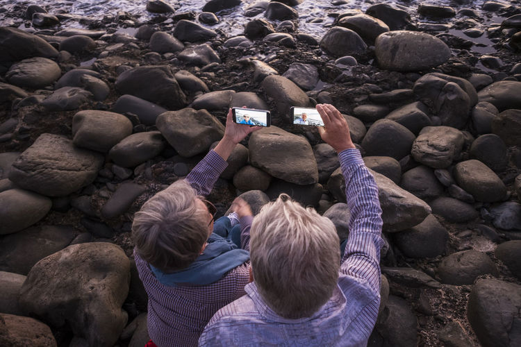 High angle view of couple taking selfie from mobile phones on rock at beach