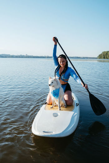 Woman with dreads paddleboarding with pet on the lake, japanese spitz dog standing on sup board