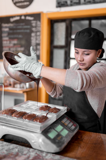 Hispanic woman in apron and gloves filling plastic mold on digital scale with liquid chocolate during work in confectionery in daytime