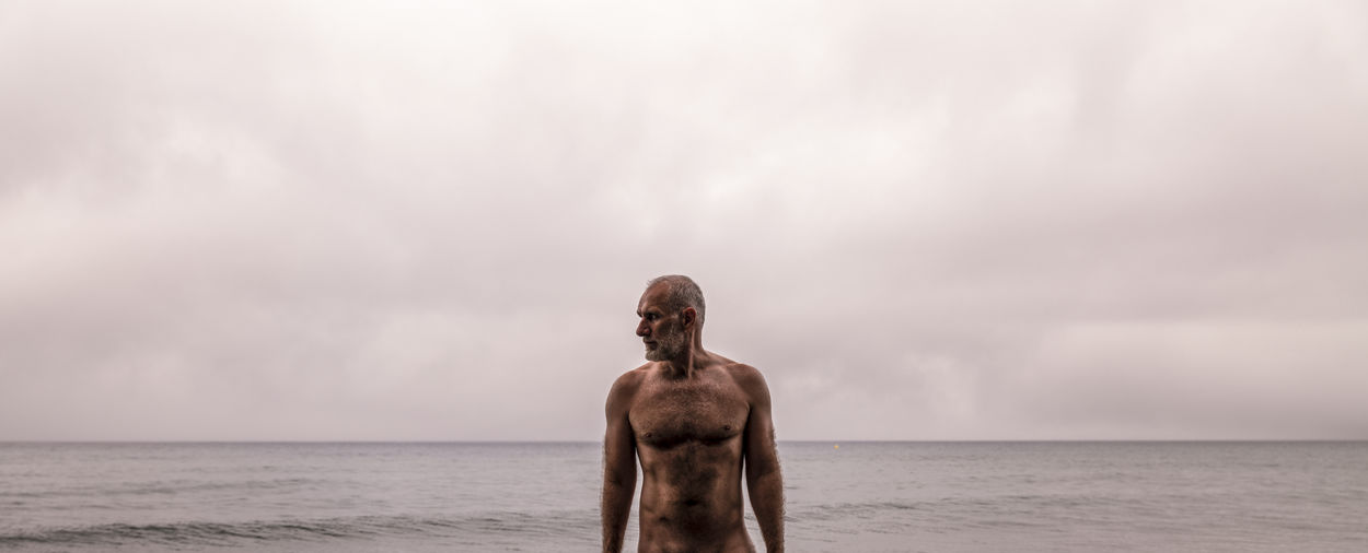 Portrait of shirtless adult man on beach against sky