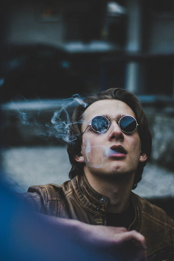 Young man wearing sunglasses and leather jacket while emitting smoke from mouth