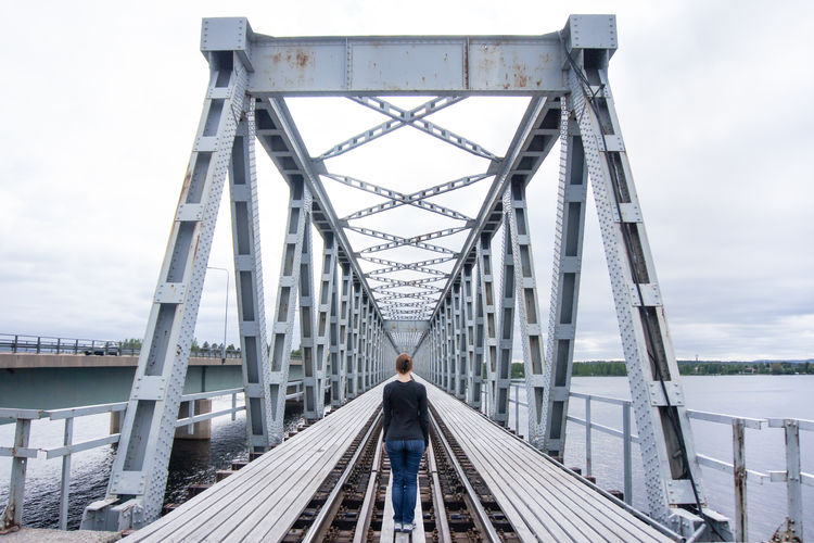 Rear view of woman standing on railway bridge over river against cloudy sky